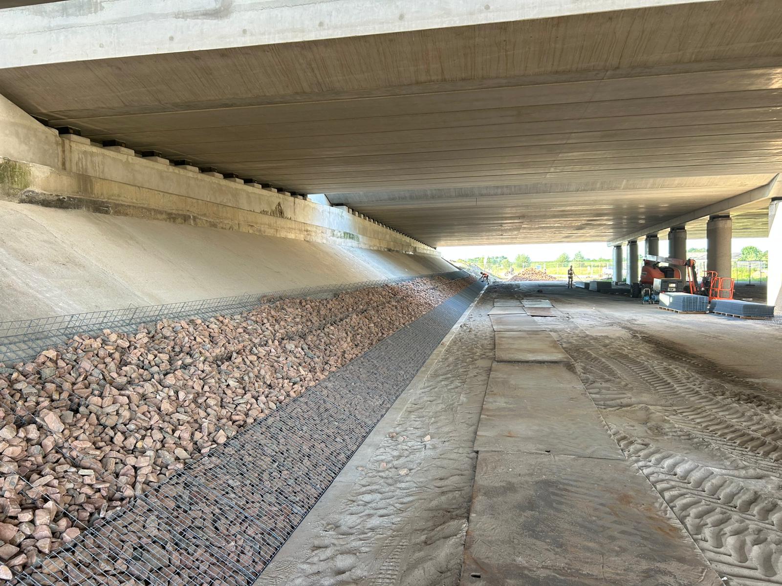 Grondkering met schanskorven onder een snelwegviaduct, gevuld met rode breuksteen